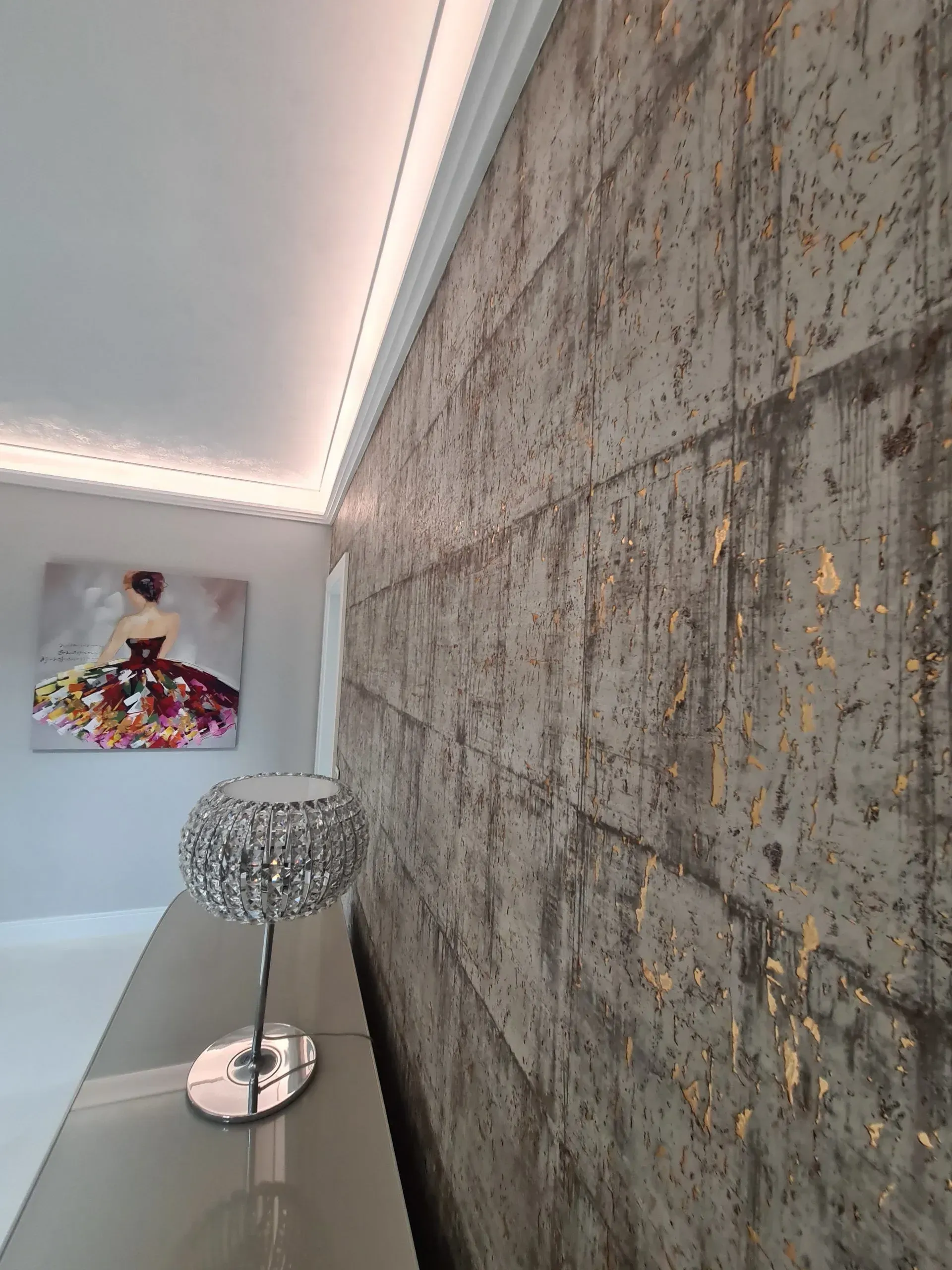 Eine strukturierte Wand in einem modernen, betonartigen Stil und goldenen Akzenten. Im Vordergrund steht eine elegante Tischlampe und im Hintergrund hängt ein farbenfrohes Gemälde. Als Maler bei Rostock bieten wir ihnen sowohl klassische als auch extravagante Wanddesigns.