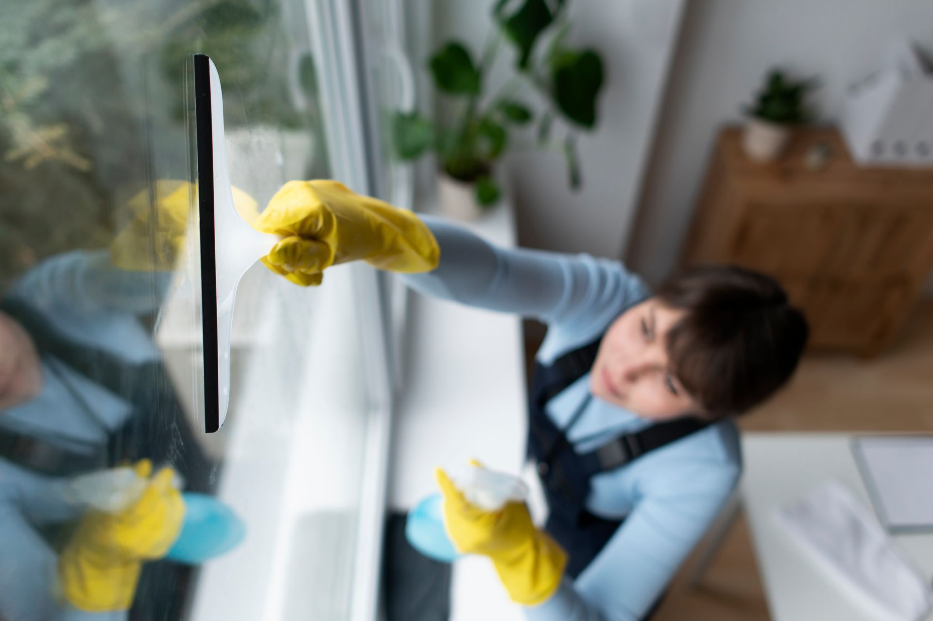 Una persona con guantes amarillos limpia una ventana con una escobilla limpiacristales en una habitación luminosa.