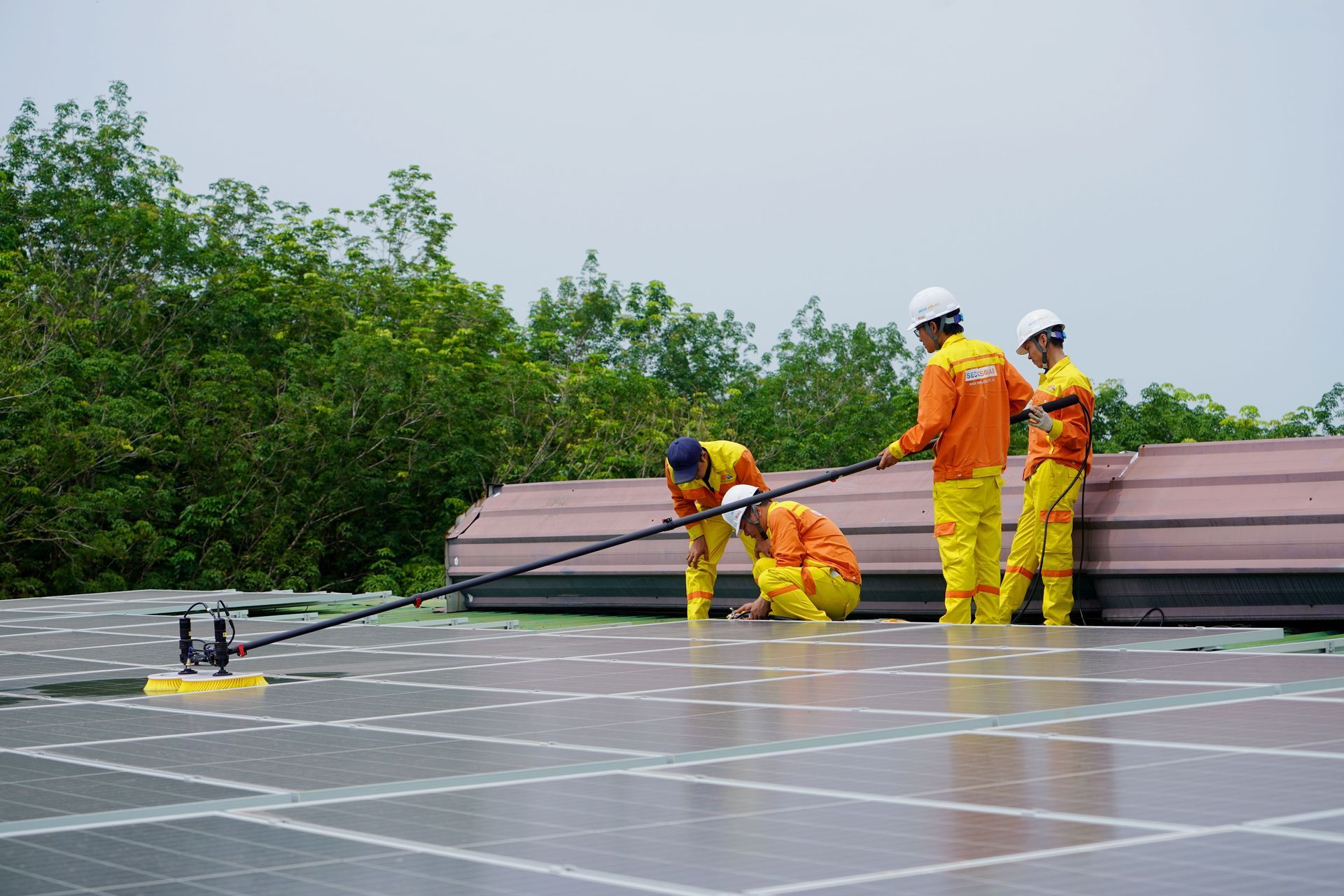 Trabajadores con trajes de seguridad naranjas inspeccionan paneles solares en una azotea cerca de materiales apilados.