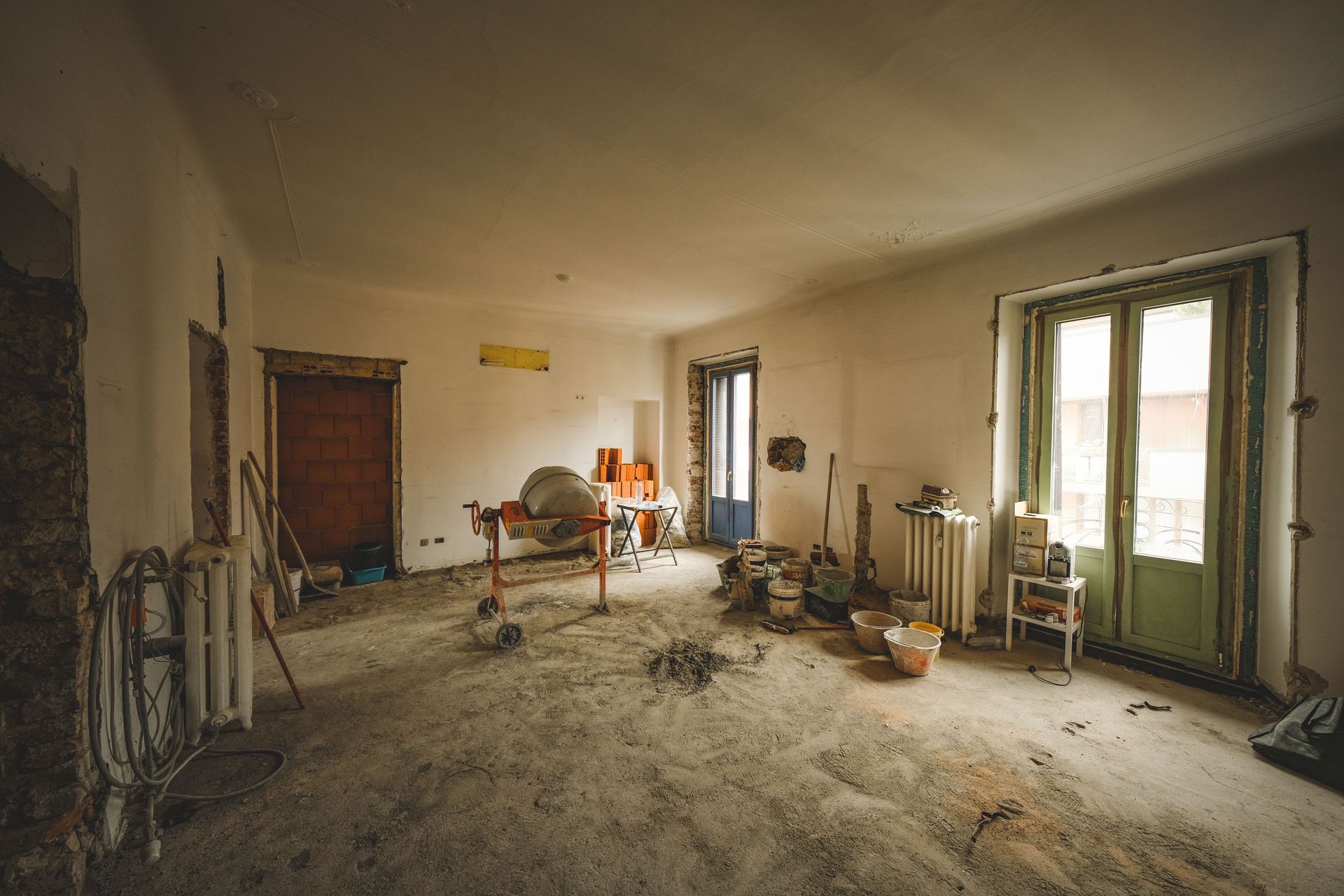 Habitación abandonada con paredes desnudas, escombros esparcidos y puertas dobles verdes que dejan entrar la luz del día.
