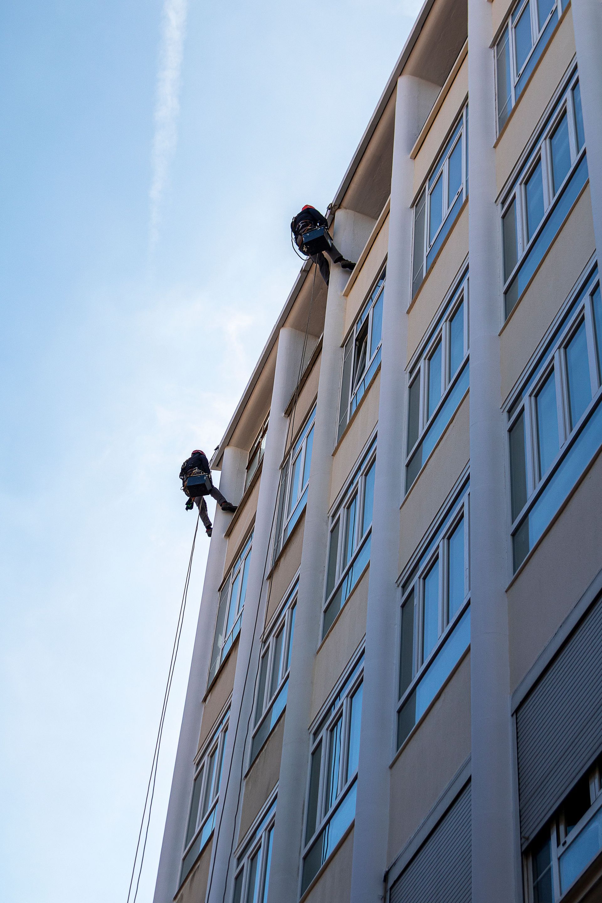 Dos limpiadores de ventanas descendiendo en rápel por la fachada de un alto edificio de oficinas contra un cielo azul