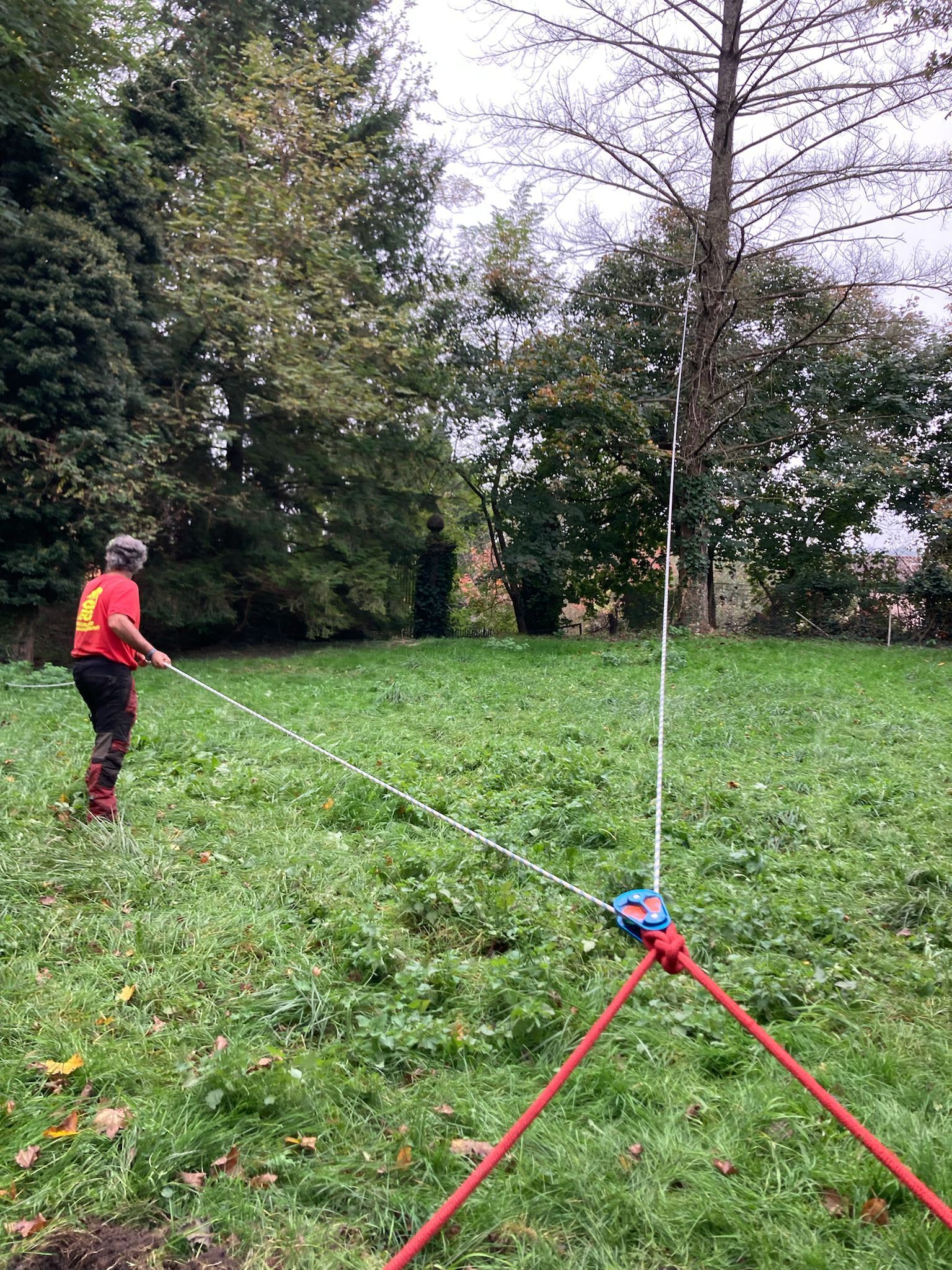 Une personne vêtue de rouge se tient debout dans un parc herbeux, utilisant une longue corde attachée à un trépied rouge.