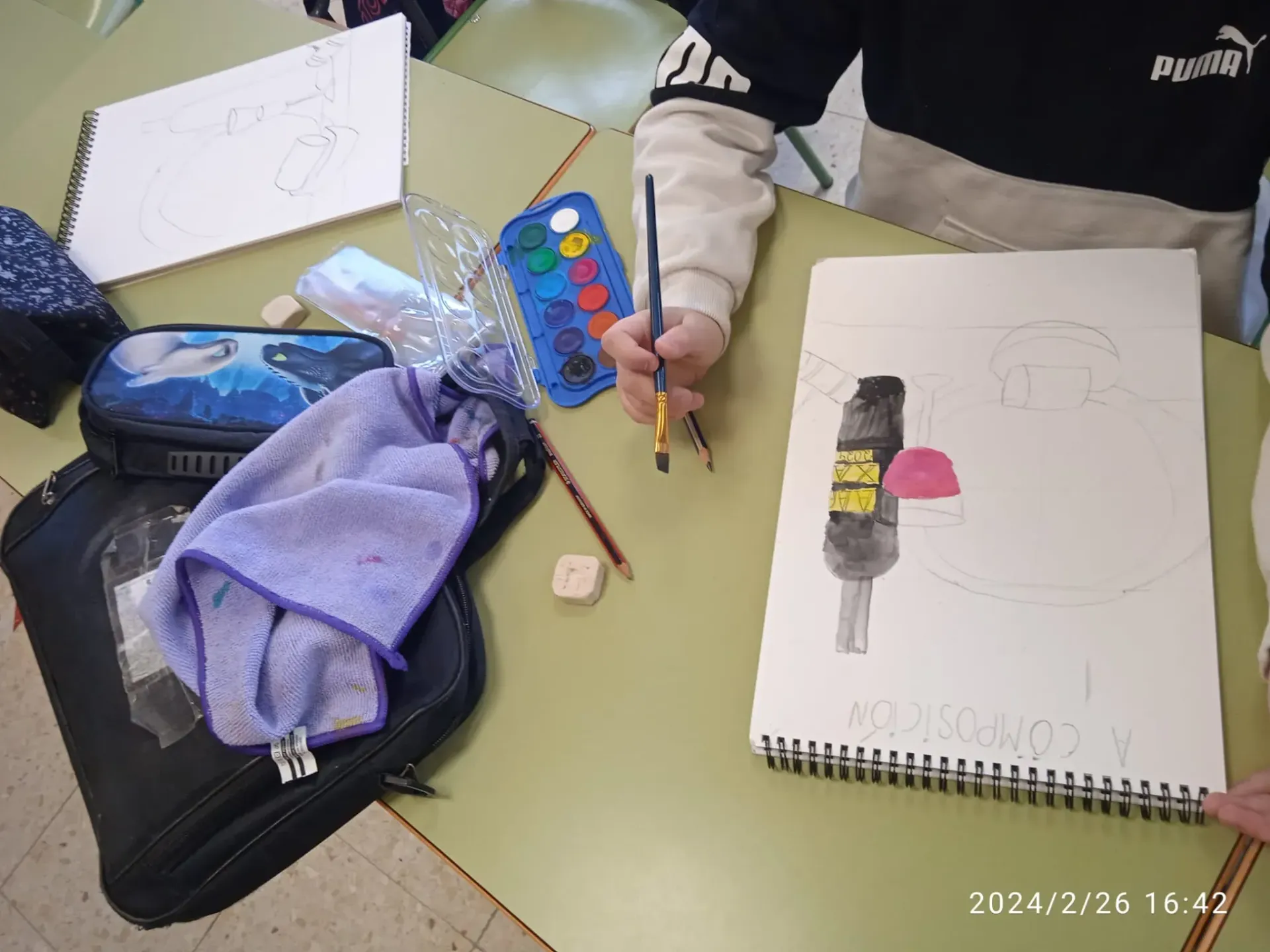 Un niño pinta en una mesa con acuarelas y un dibujo en papel junto a sus materiales de arte.