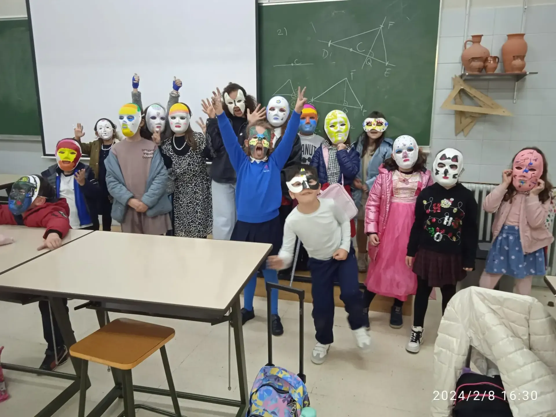 Niños disfrazados y con máscaras están de pie en un aula con pupitres y una pizarra, algunos levantan la mano.