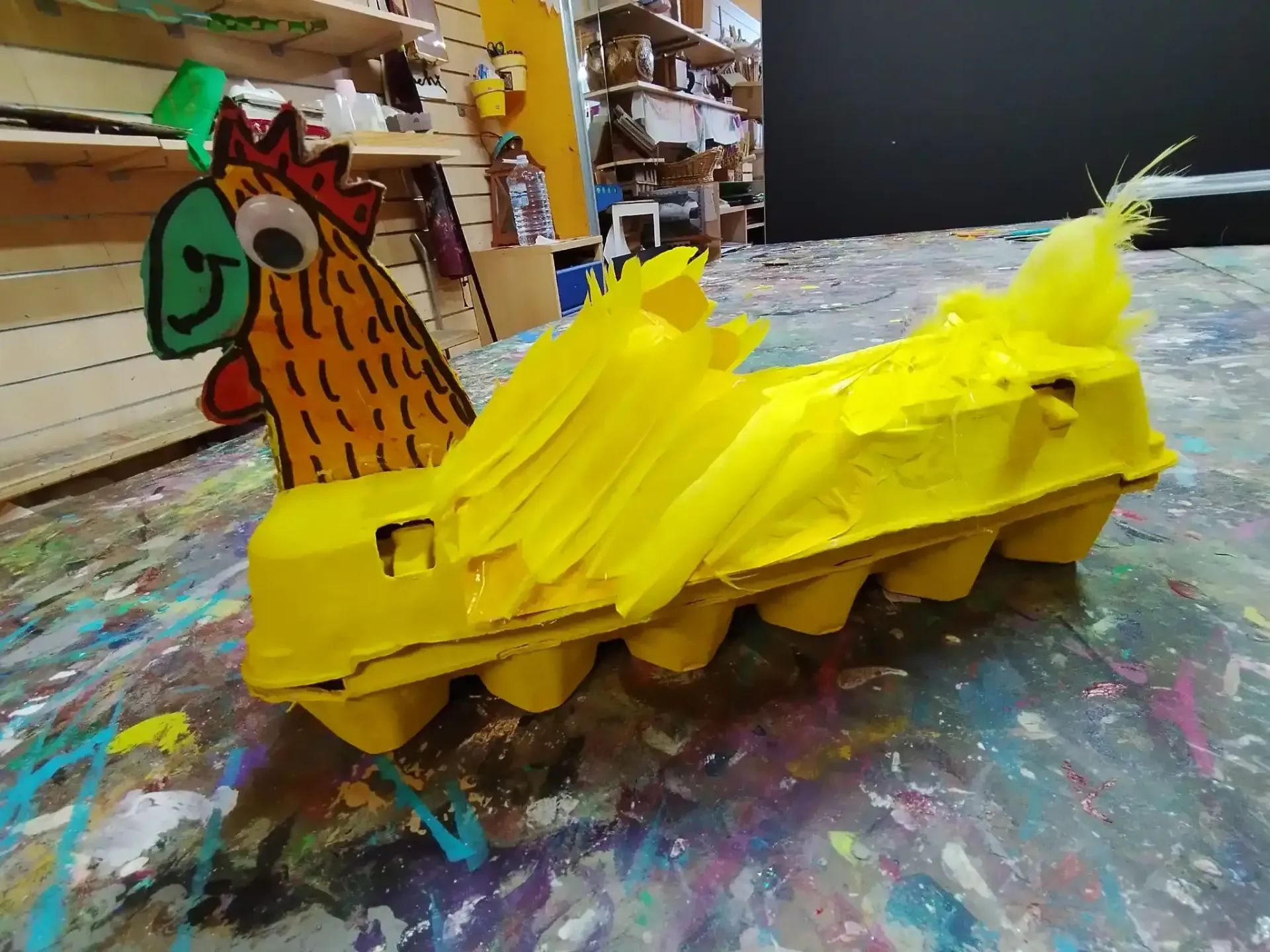 Maqueta amarilla con una cabeza de pollo pintada sobre una mesa salpicada de pintura en un aula de arte.