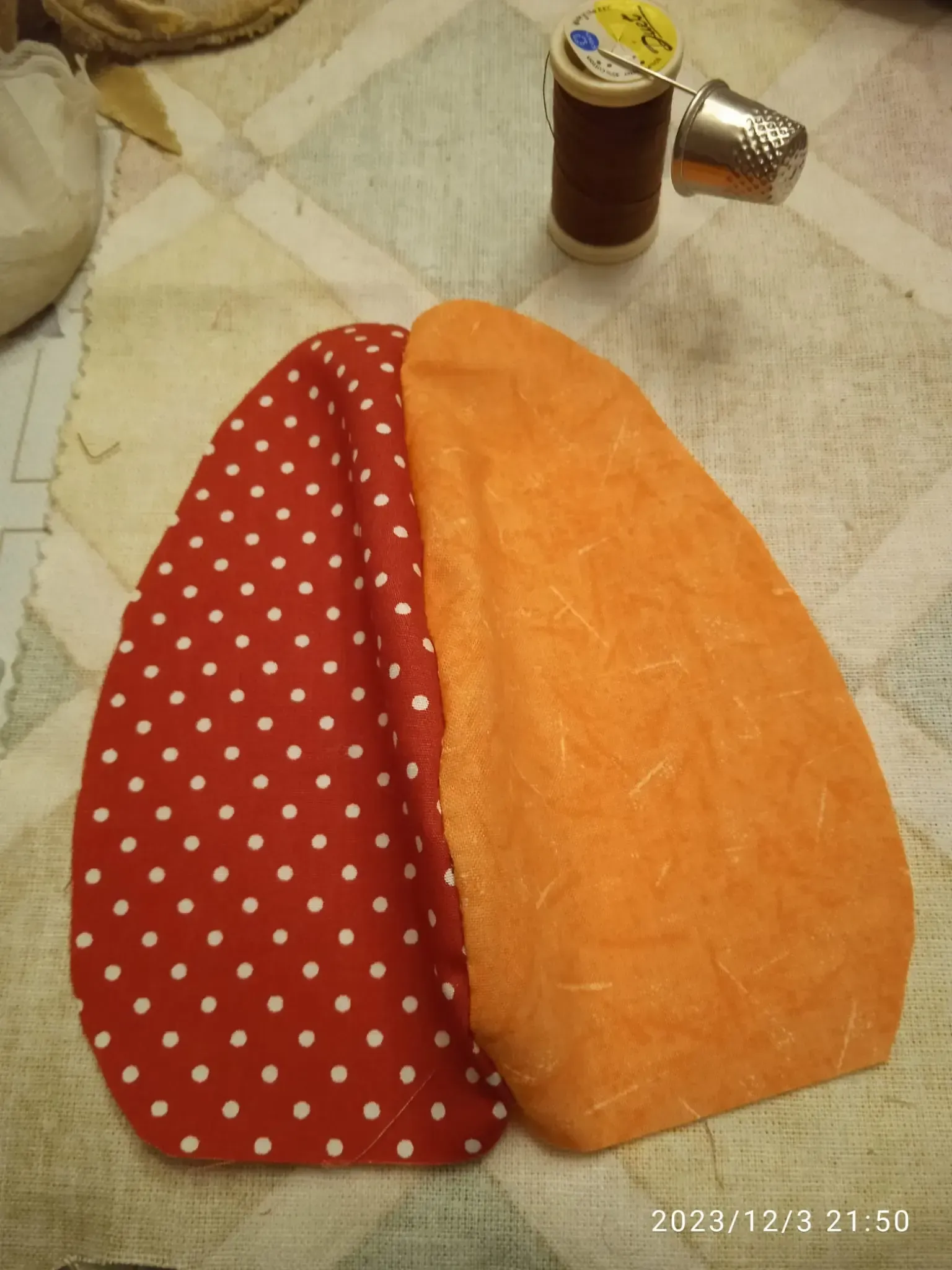 Trozos de tela roja y naranja sobre una cama, con un carrete de hilo marrón y un dedal de metal cerca.