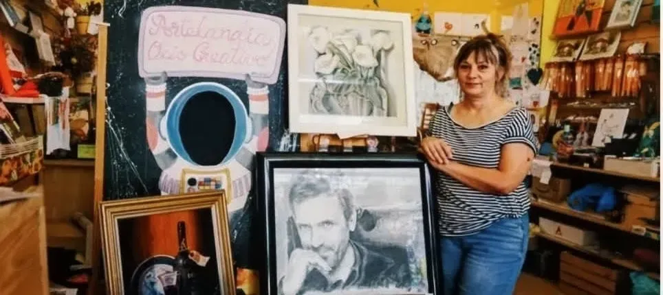 Artista de pie en un estudio desordenado junto a retratos enmarcados y bocetos en la pared.