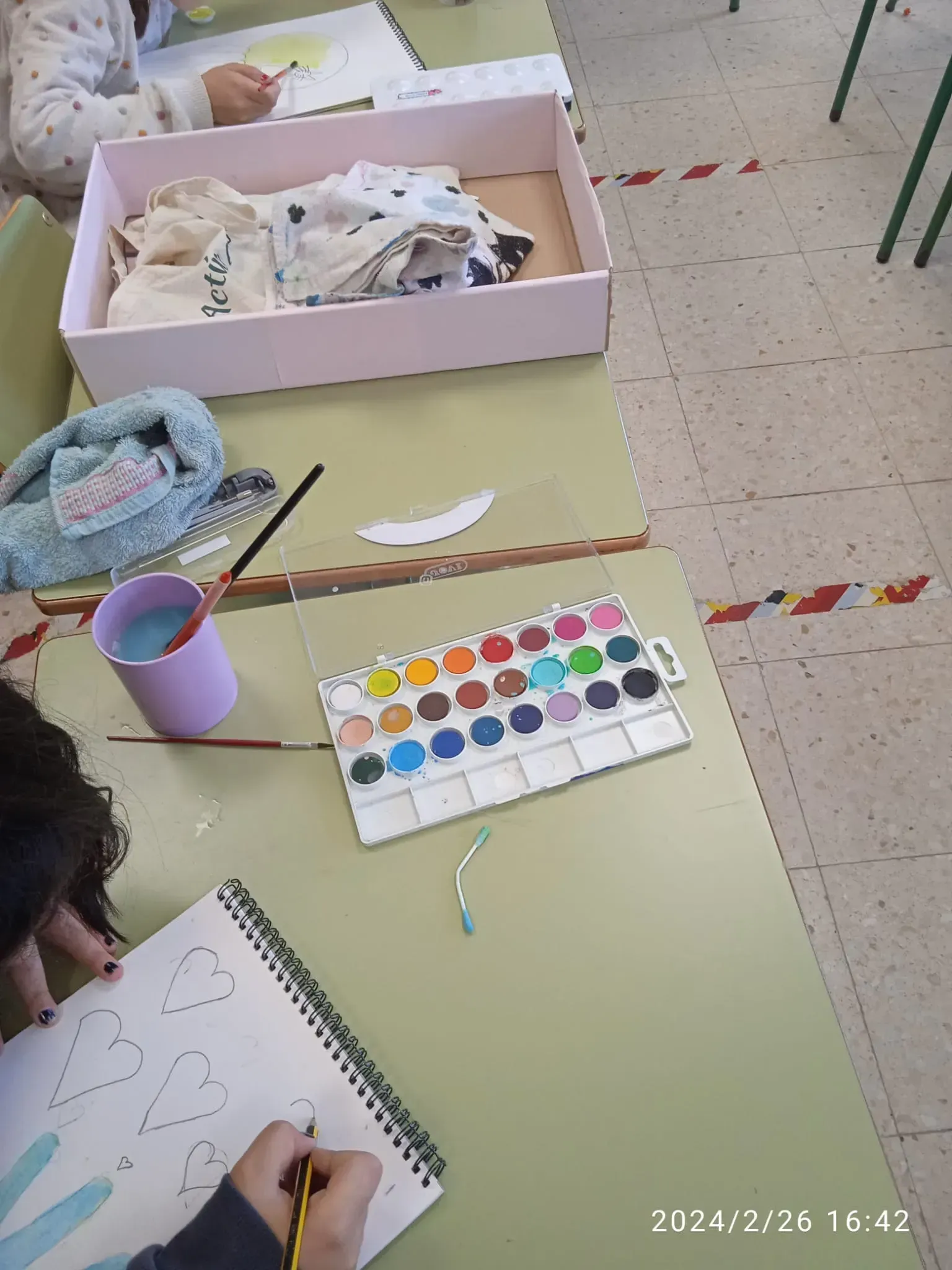 Una persona dibuja en una mesa de aula con acuarelas, pinceles y materiales de arte a su alcance.