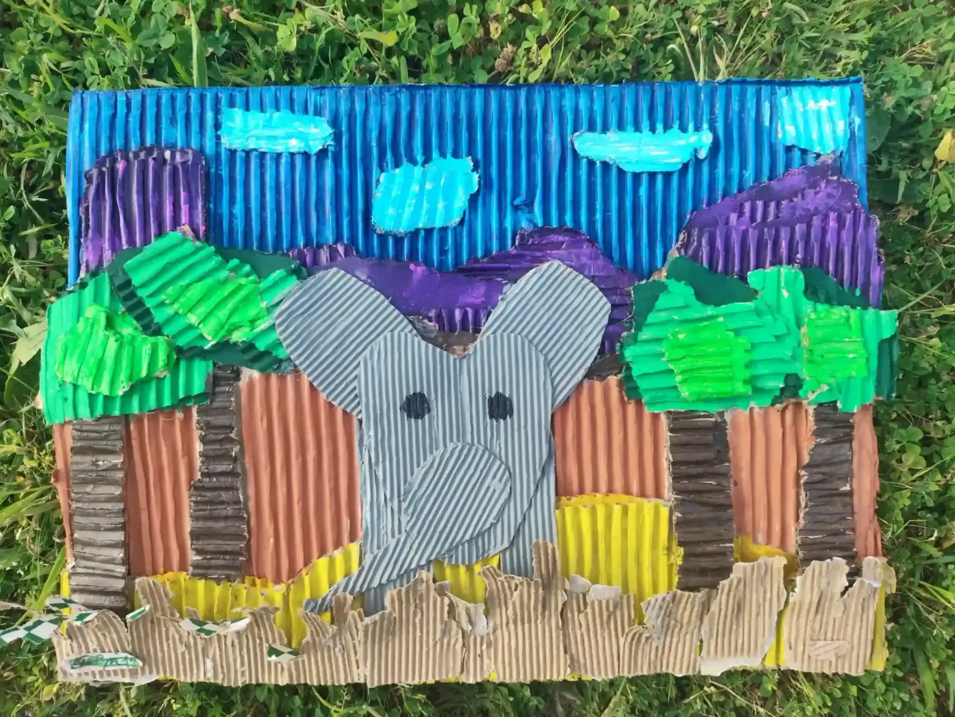Dibujo infantil y colorido de un elefante gris entre chozas, con árboles verdes, montañas moradas y cielo azul.