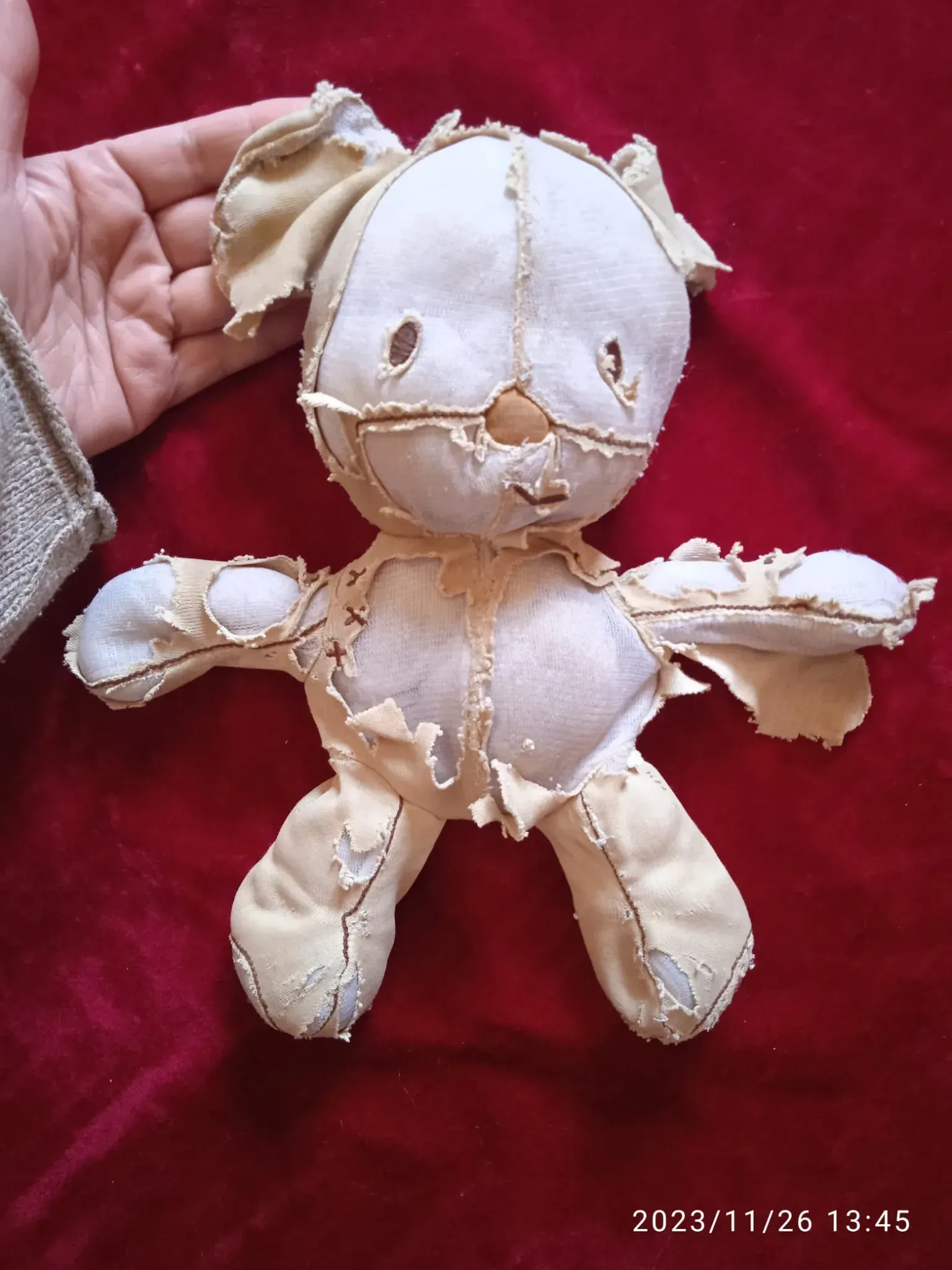 Mano sosteniendo una muñeca de trapo beige desgastada con costuras cosidas sobre un fondo de tela roja.