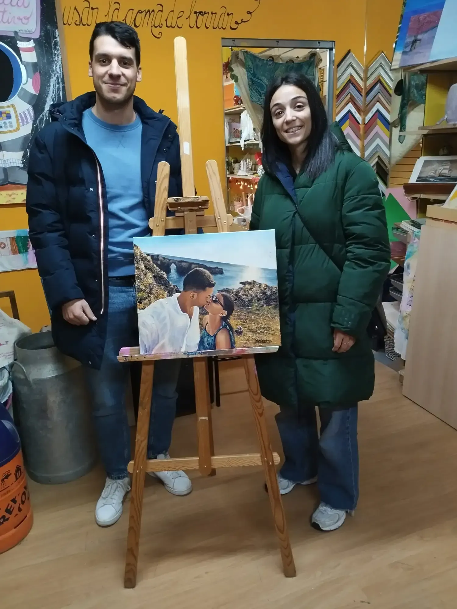 Dos personas de pie junto a un caballete que exhibe un cuadro de paisaje en un colorido estudio de arte.
