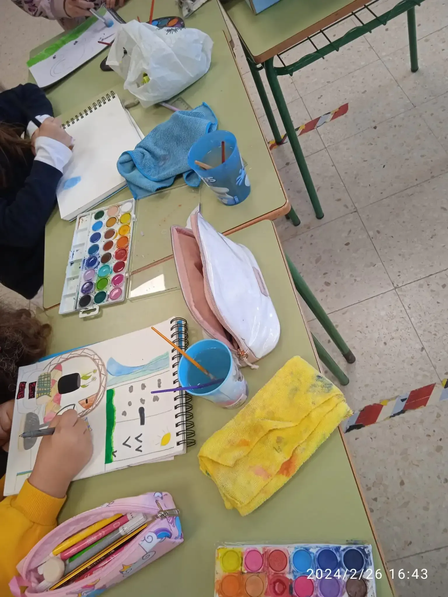 Alumnos pintando en sus pupitres con paletas de acuarela, pinceles y estuches de lápices en un aula.