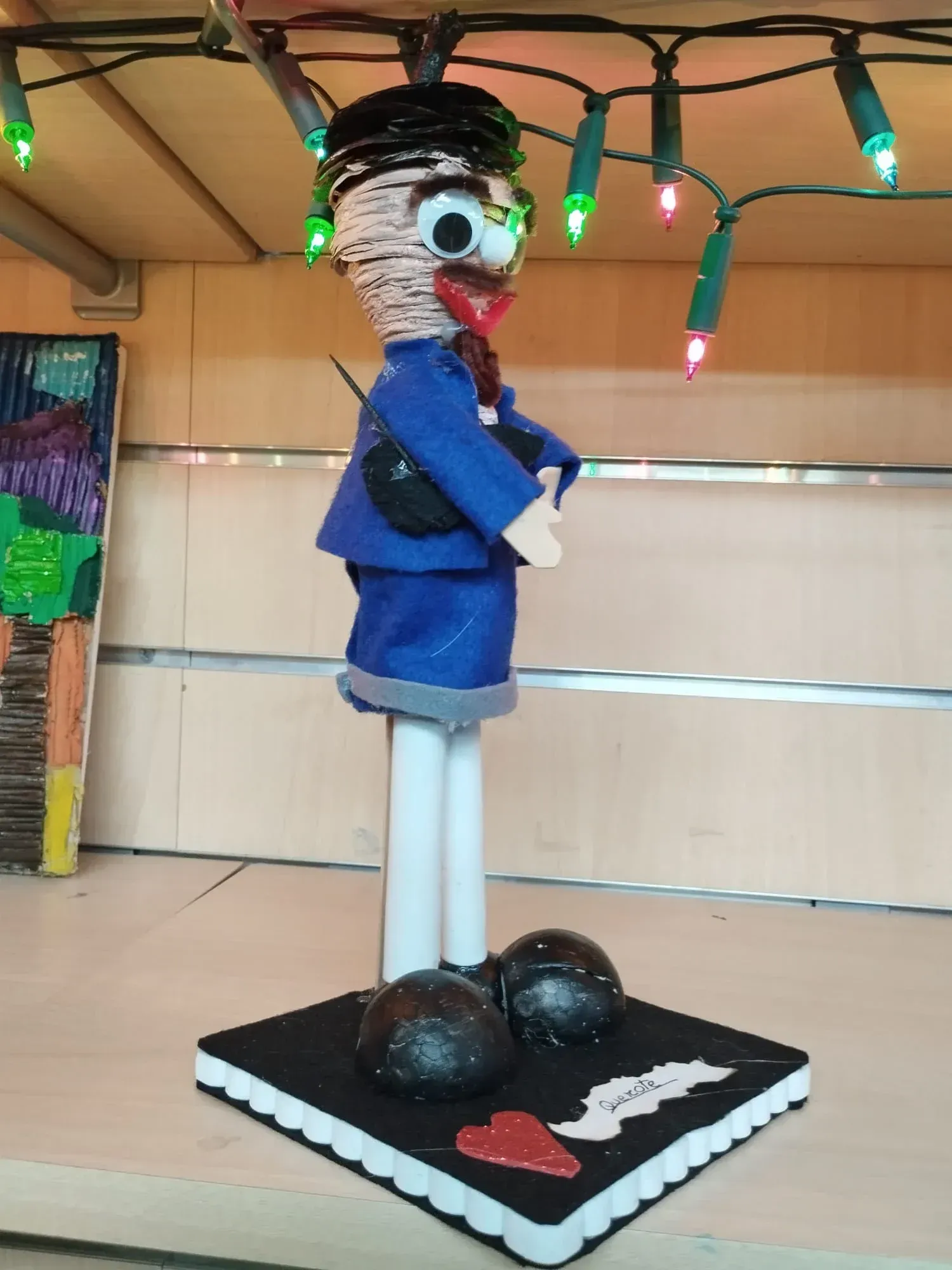 Pequeña figura artesanal con abrigo azul sobre una base negra con luces en la parte superior.