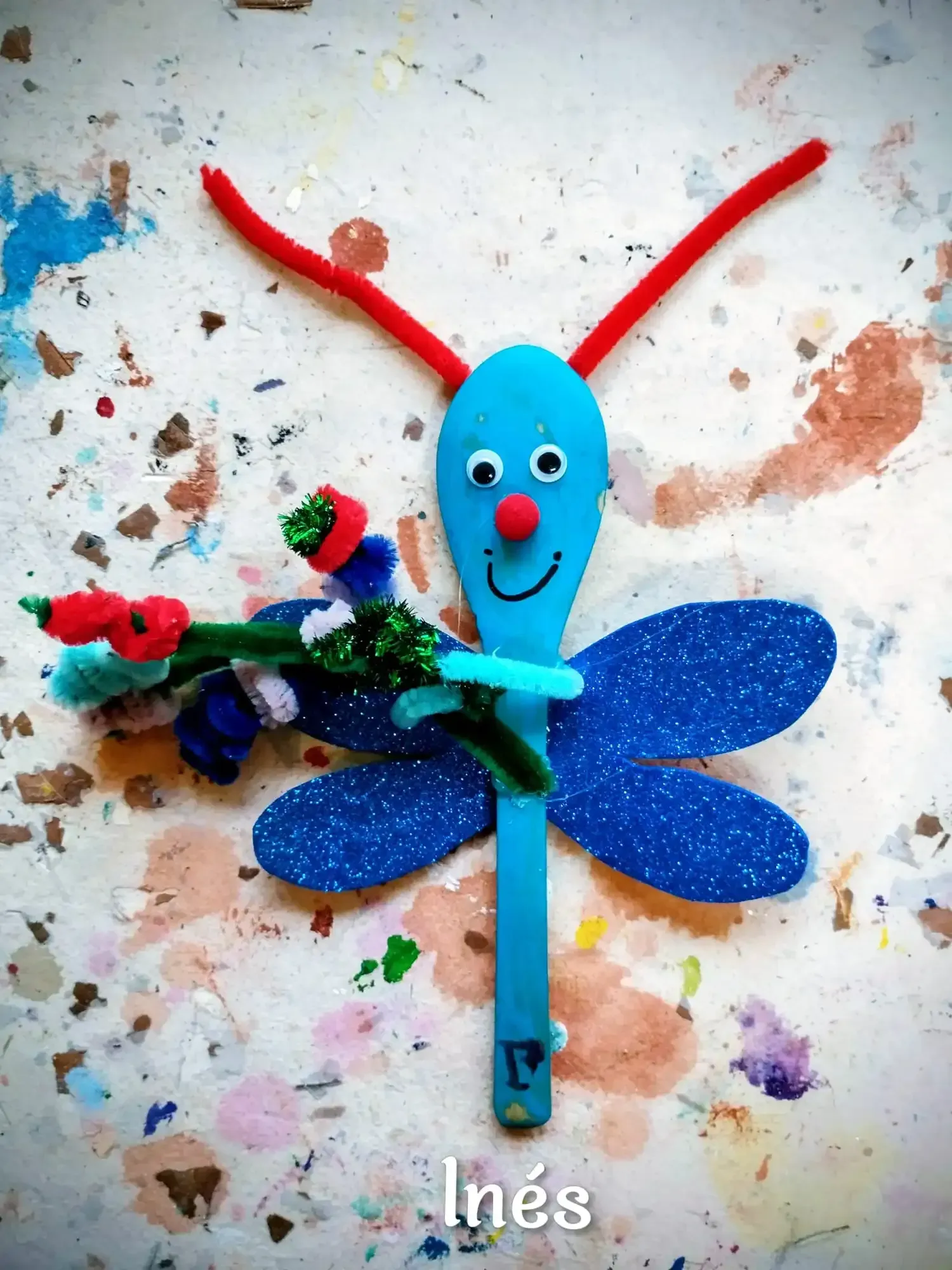 Insecto artesanal azul con antenas rojas y alas brillantes sobre un fondo salpicado de pintura.