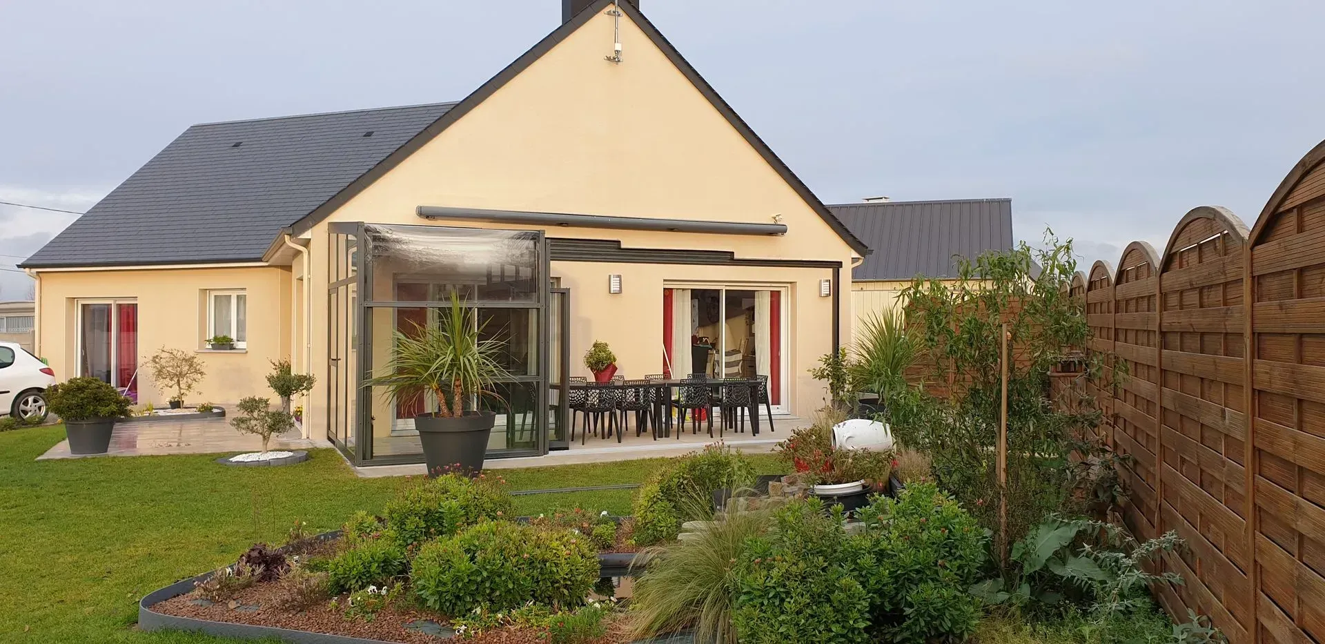 Maison beige de plain-pied avec toit foncé, terrasse et jardin paysager sous un ciel nuageux