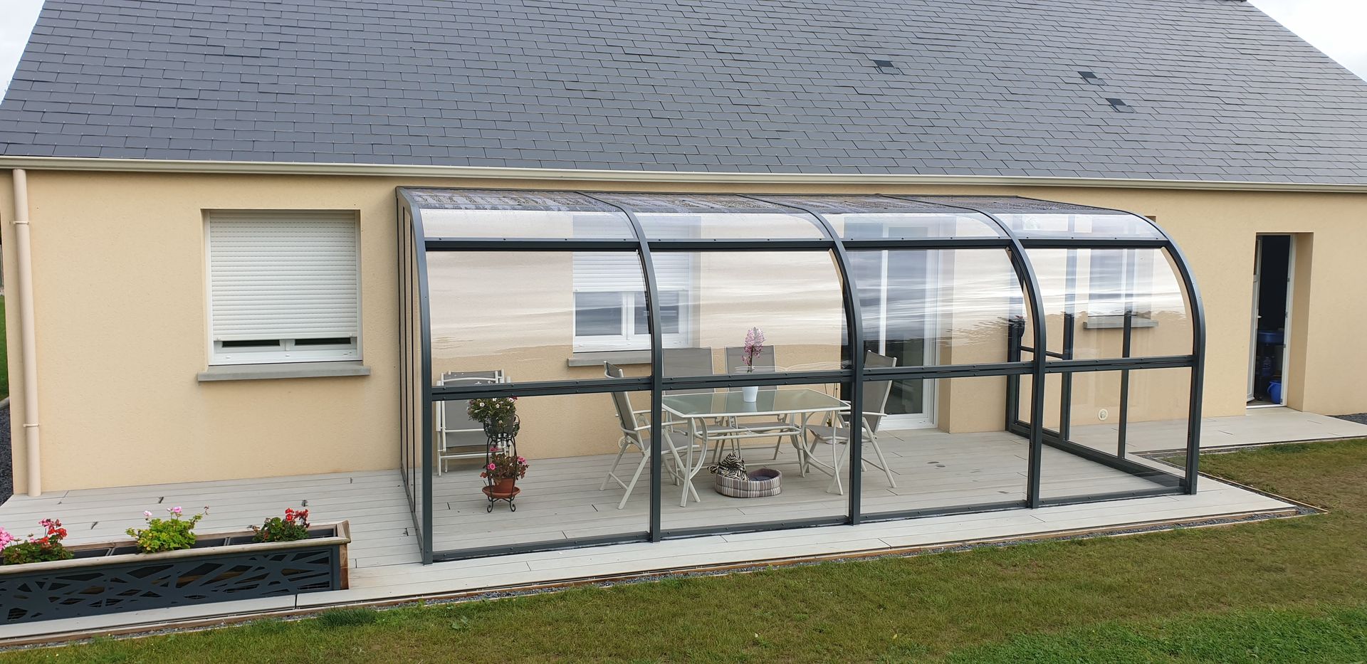 Patio vitré attenant à une maison beige, avec des places assises en extérieur et une pelouse à l’avant