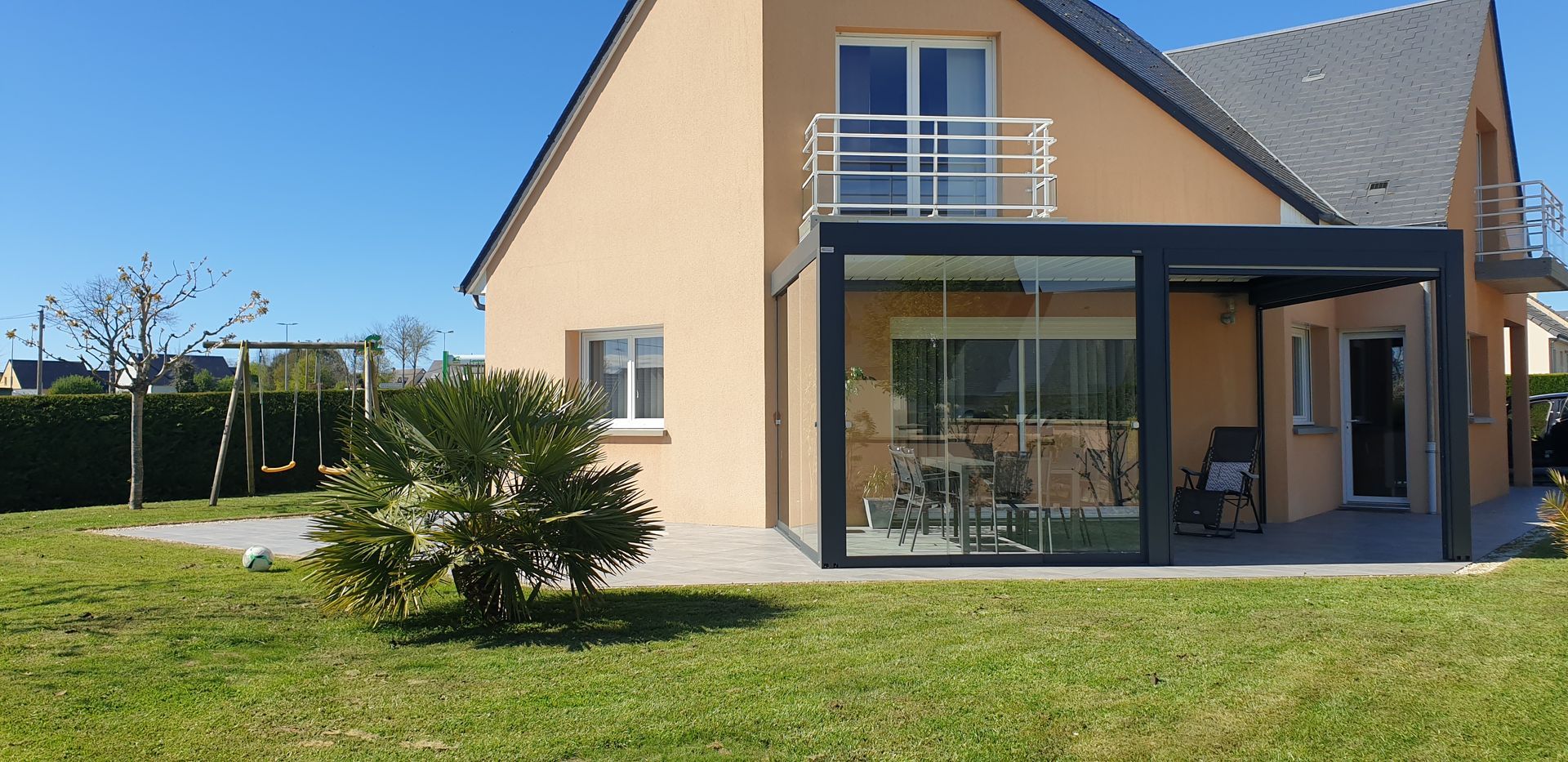 Maison beige moderne avec balcon, grandes fenêtres et pelouse verte sous un ciel bleu azur.