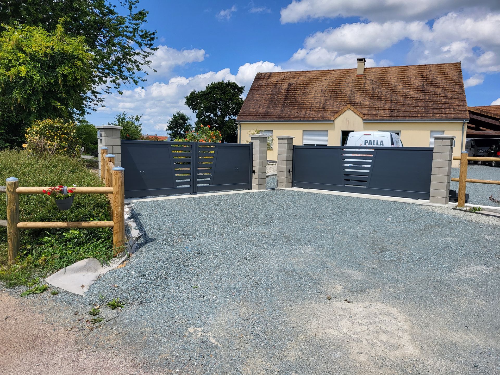 Allée en gravier menant à une maison avec un portail gris foncé, une clôture en bois et des arbres sous un ciel bleu.