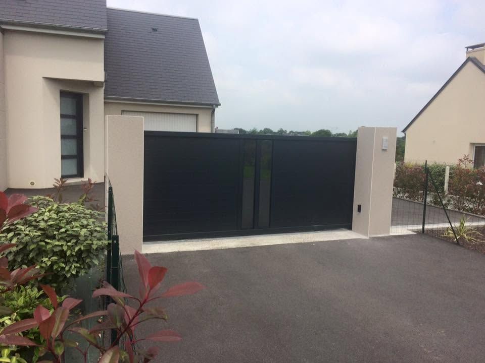 Entrée de maison moderne avec portail coulissant noir et allée carrossable, bordée de murs et d'arbustes de couleur claire.