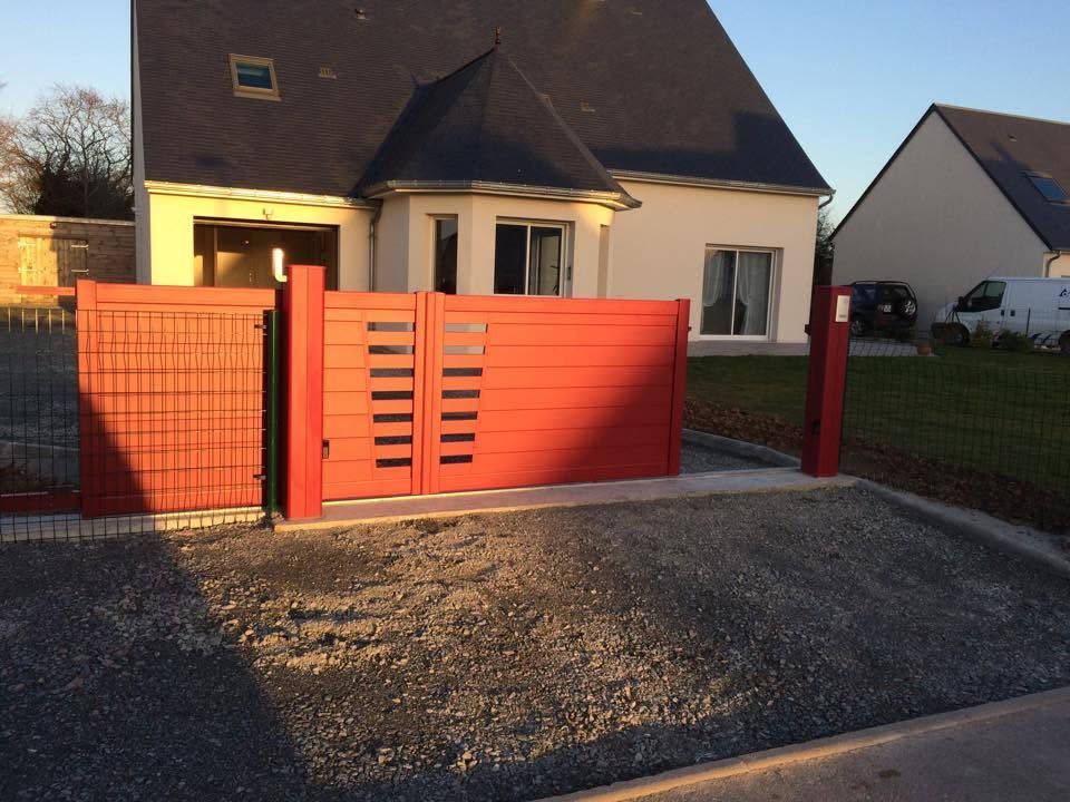 Portail et clôture rouges devant une maison couleur crème, au bord d'une allée de gravier.