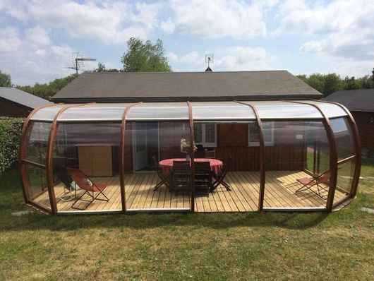 Terrasse circulaire fermée par des moustiquaires, avec terrasse en bois, table, chaises et barbecue dans un jardin