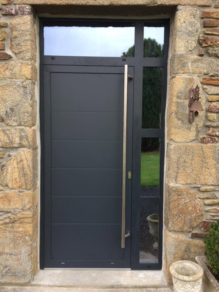 Porte d'entrée gris foncé encastrée dans un mur en pierre, avec un panneau latéral en verre étroit et une poignée verticale.