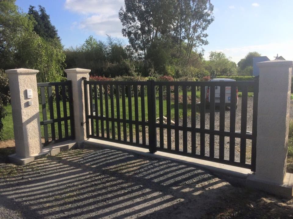 Portail d'allée en métal noir entre des piliers de pierre, projetant des ombres rayées au soleil.