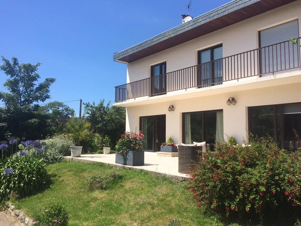 Maison à deux étages avec balcon et jardin, façade blanche, sous un ciel bleu azur.