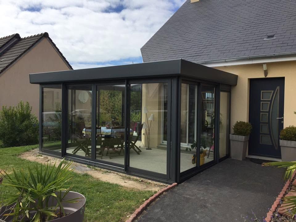 Extension moderne de type véranda vitrée attenante à la maison, avec une structure sombre et une terrasse pavée.