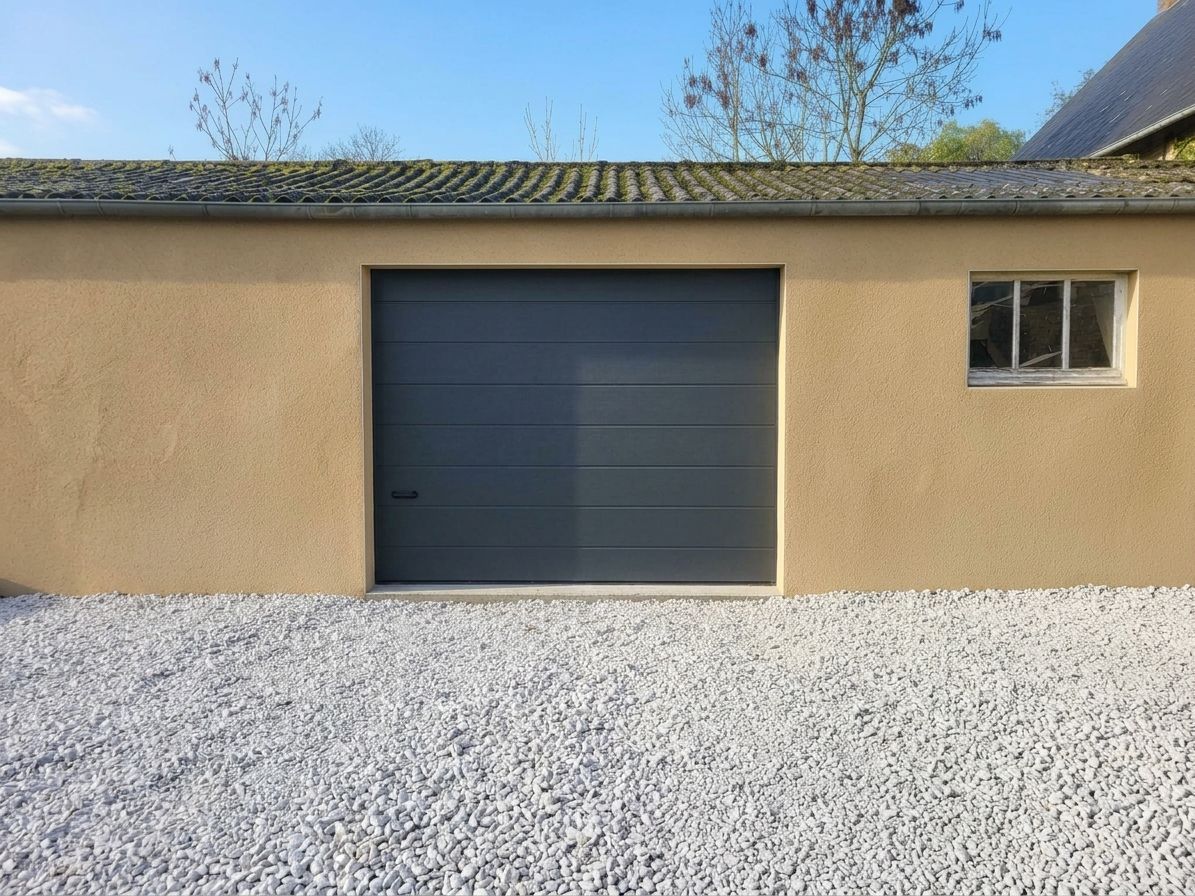 Garage beige avec une porte bleu foncé et une petite fenêtre, donnant sur une cour gravillonnée.