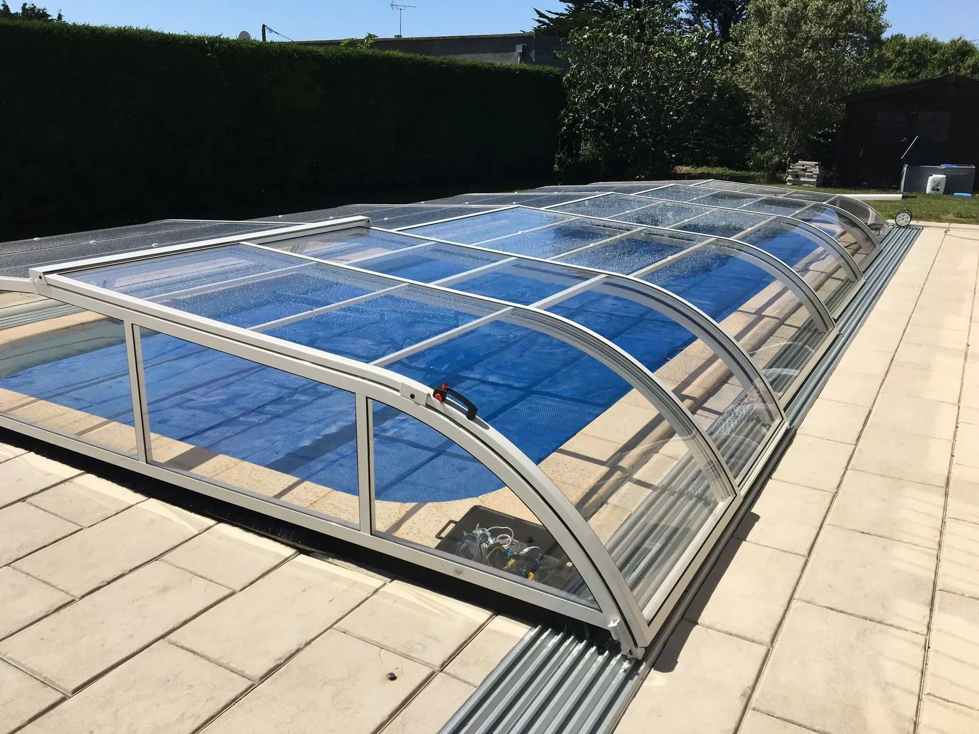 Couverture de piscine bleue sous une structure métallique incurvée, à côté d'une terrasse pavée et de haies.