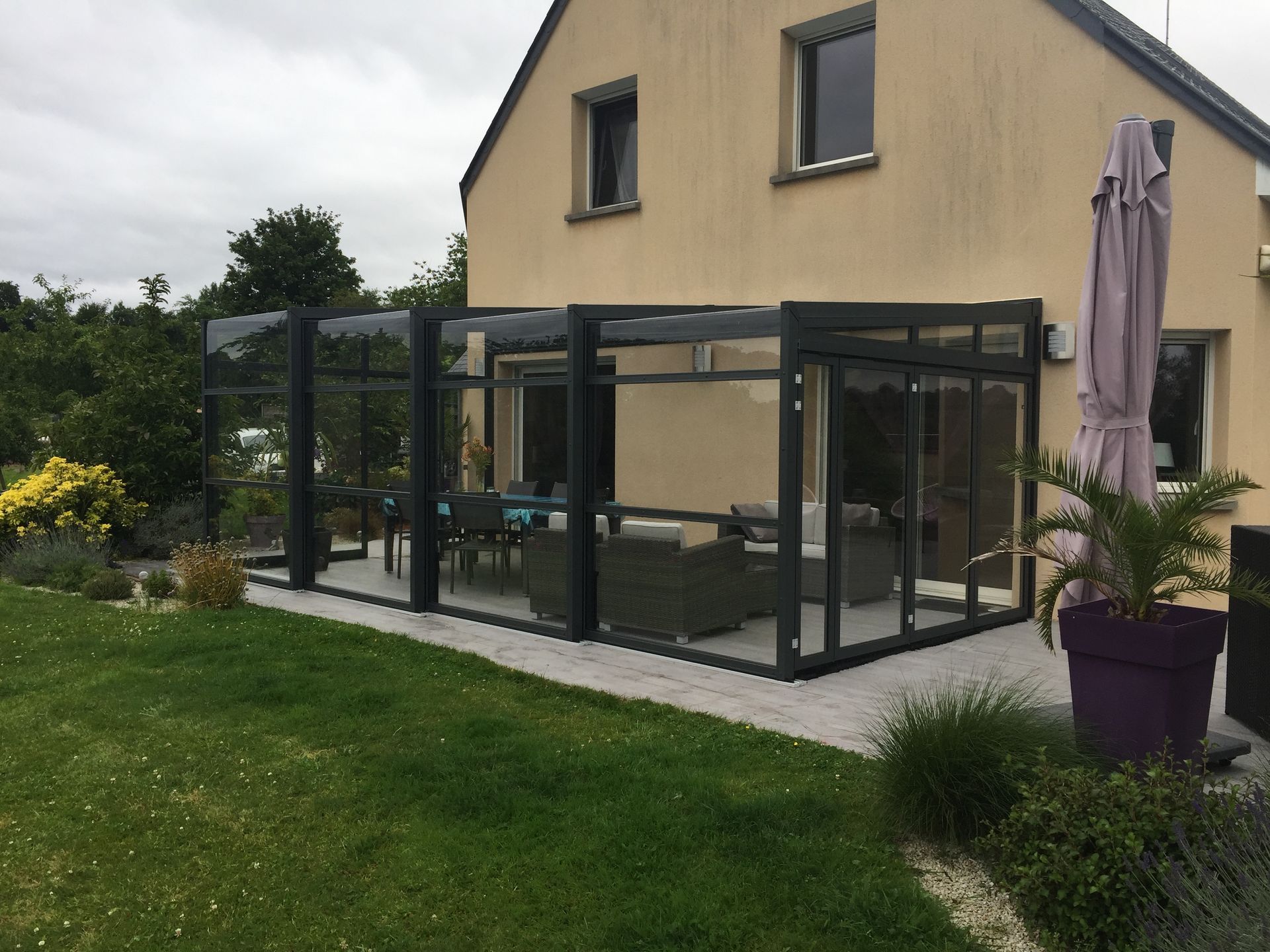 Patio vitré attenant à une maison beige, avec un parasol fermé et des plantes en pot sur la terrasse.