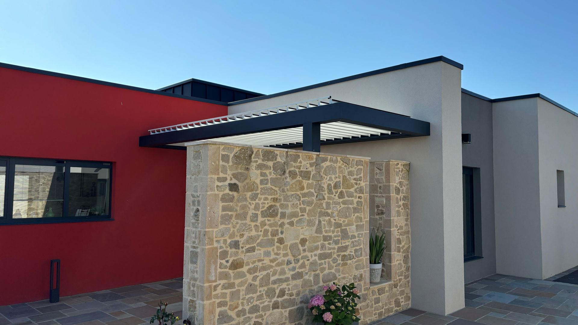 Entrée d'un bâtiment moderne avec mur en pierre, façade rouge et grise, et petite porte couverte.