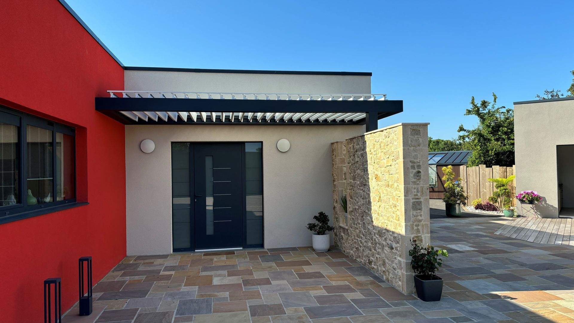 Entrée de maison moderne avec murs rouges et beiges, porte d'entrée foncée, terrasse en pierre et plantes en pot
