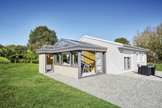 Maison moderne blanche avec véranda vitrée, allée en gravier gris et pelouse verte sous un ciel bleu