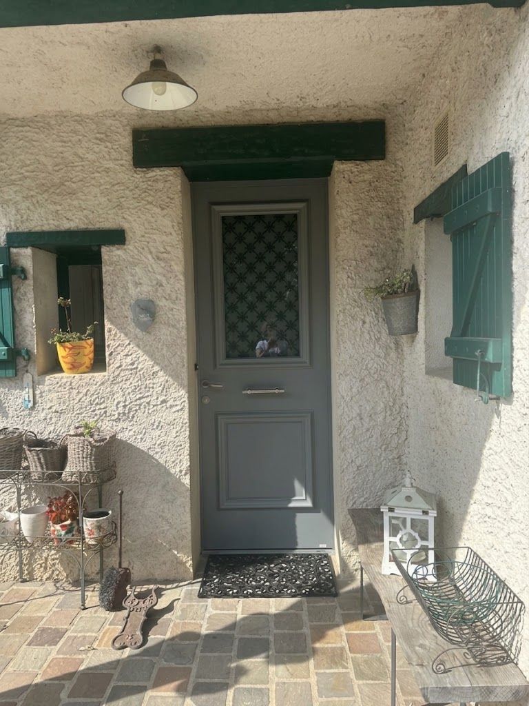 Porche d’entrée avec une porte grise, des volets verts, une lanterne murale et des plantes en pot sur un seuil carrelé.