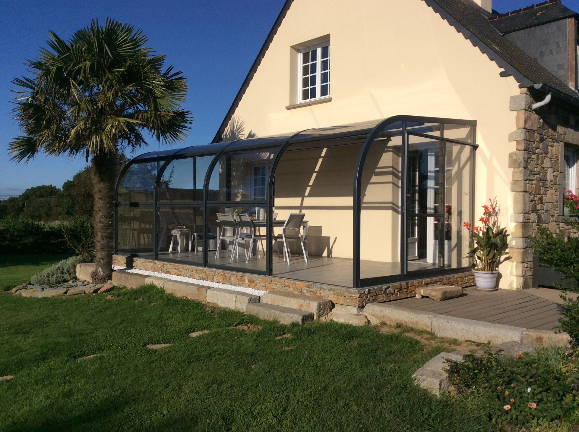 Maison baignée de soleil avec véranda vitrée, terrasse en pierre et palmier bordant une pelouse verdoyante.