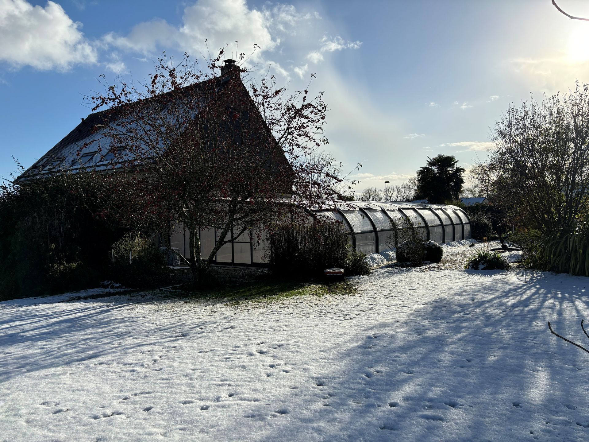 Ferme enneigée avec serre sous un ciel d’hiver lumineux, de longues ombres s’étirant dans la cour