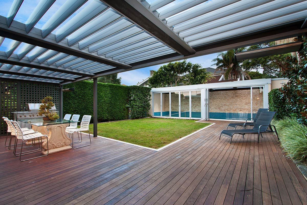 Terrasse couverte en bois avec sièges extérieurs, pelouse verte et portes d'entrée vitrées.