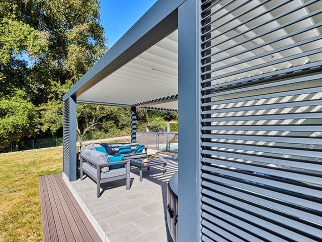 Patio couvert avec sièges gris, coussins turquoise et terrasse en bois jouxtant un mur grillagé.