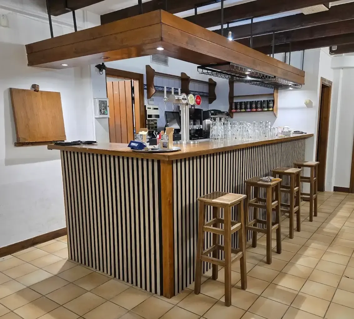 Barra de madera con taburetes. Tiradores de grifo y cristalería visibles. Tonos marrones y tostados.