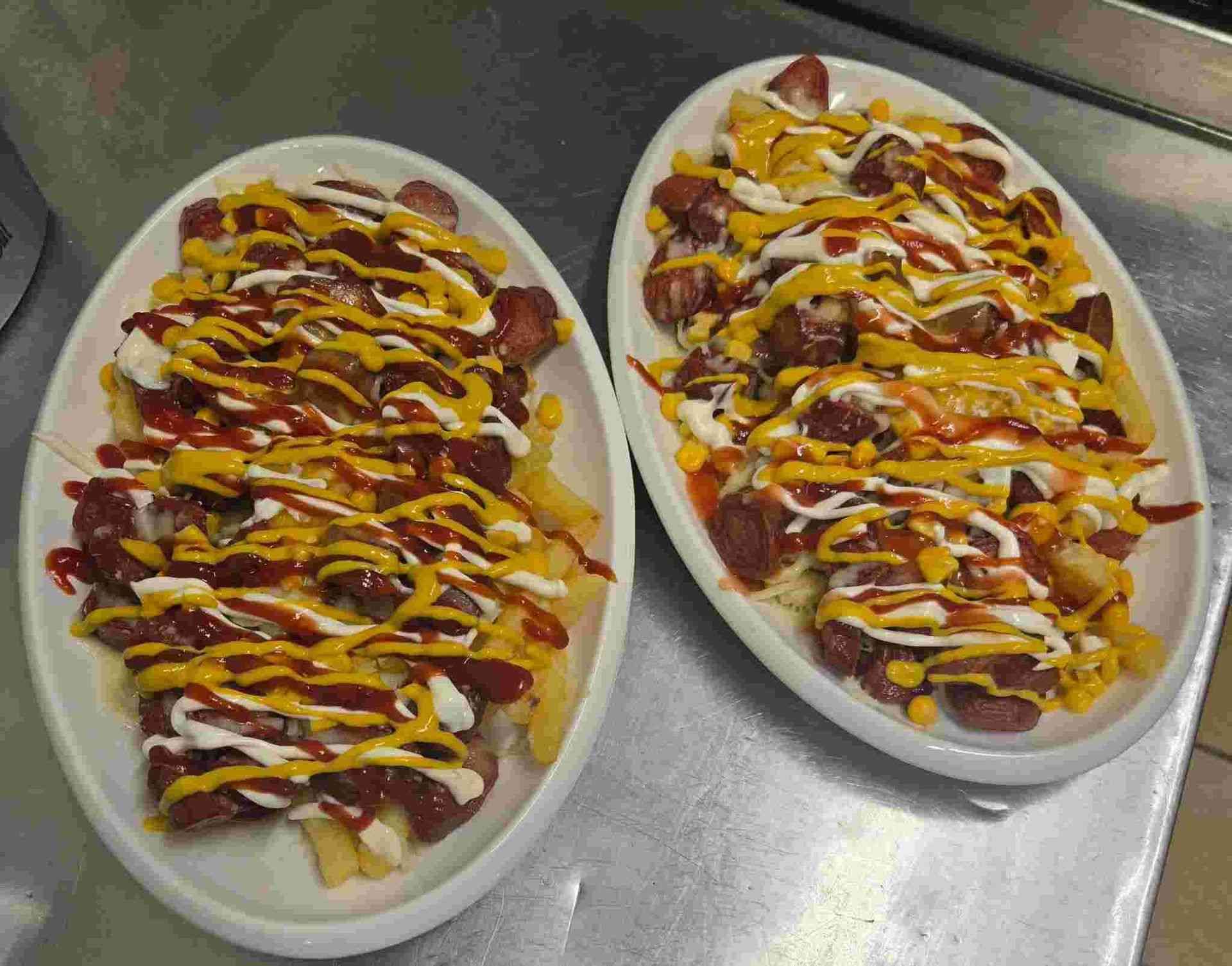 Dos platos ovalados de papas fritas cargadas, cubiertas con salchicha, ketchup, mostaza y salsa blanca.