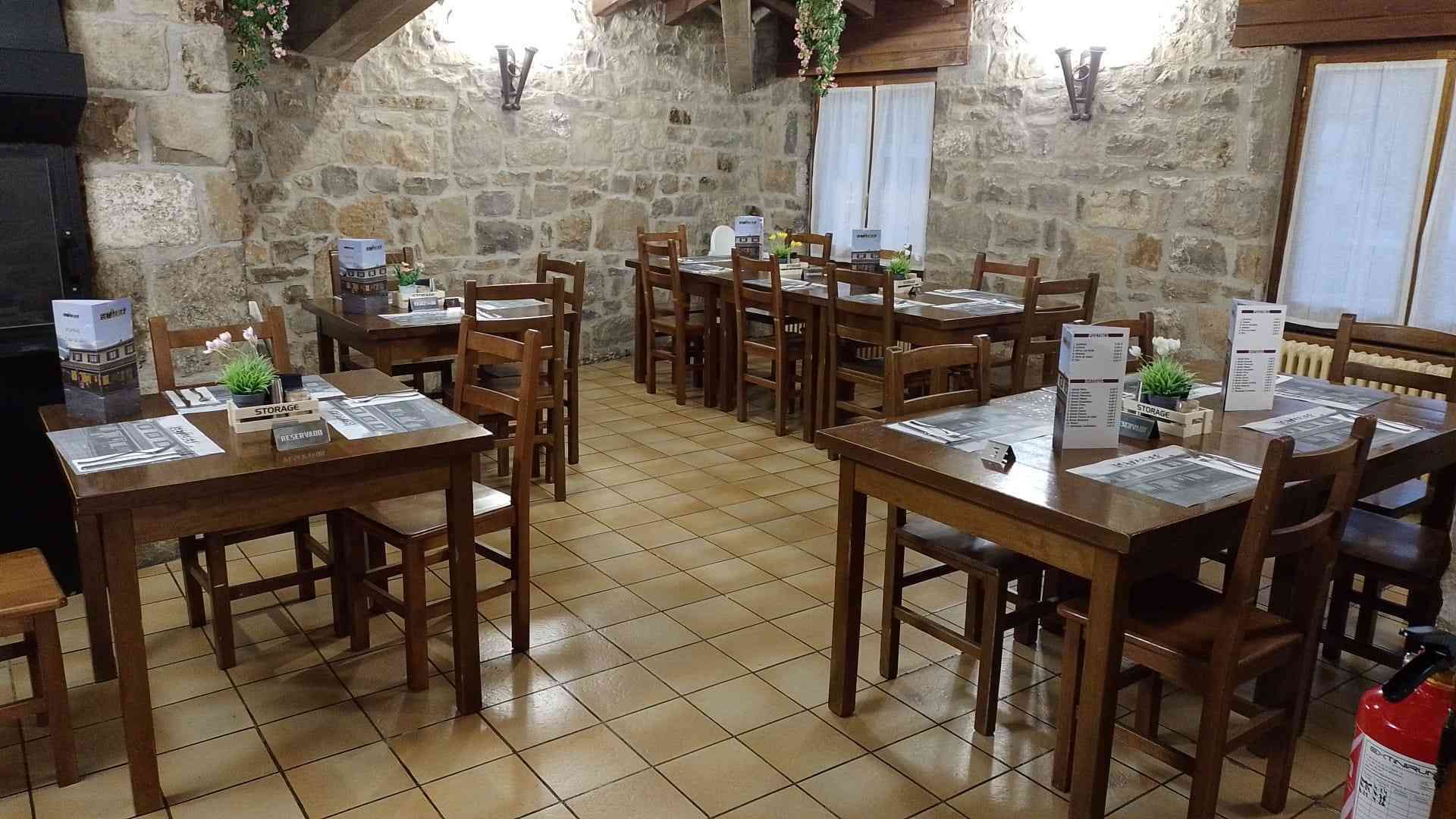 Interior de restaurante con mesas y sillas de madera, paredes de piedra y suelo de baldosas.