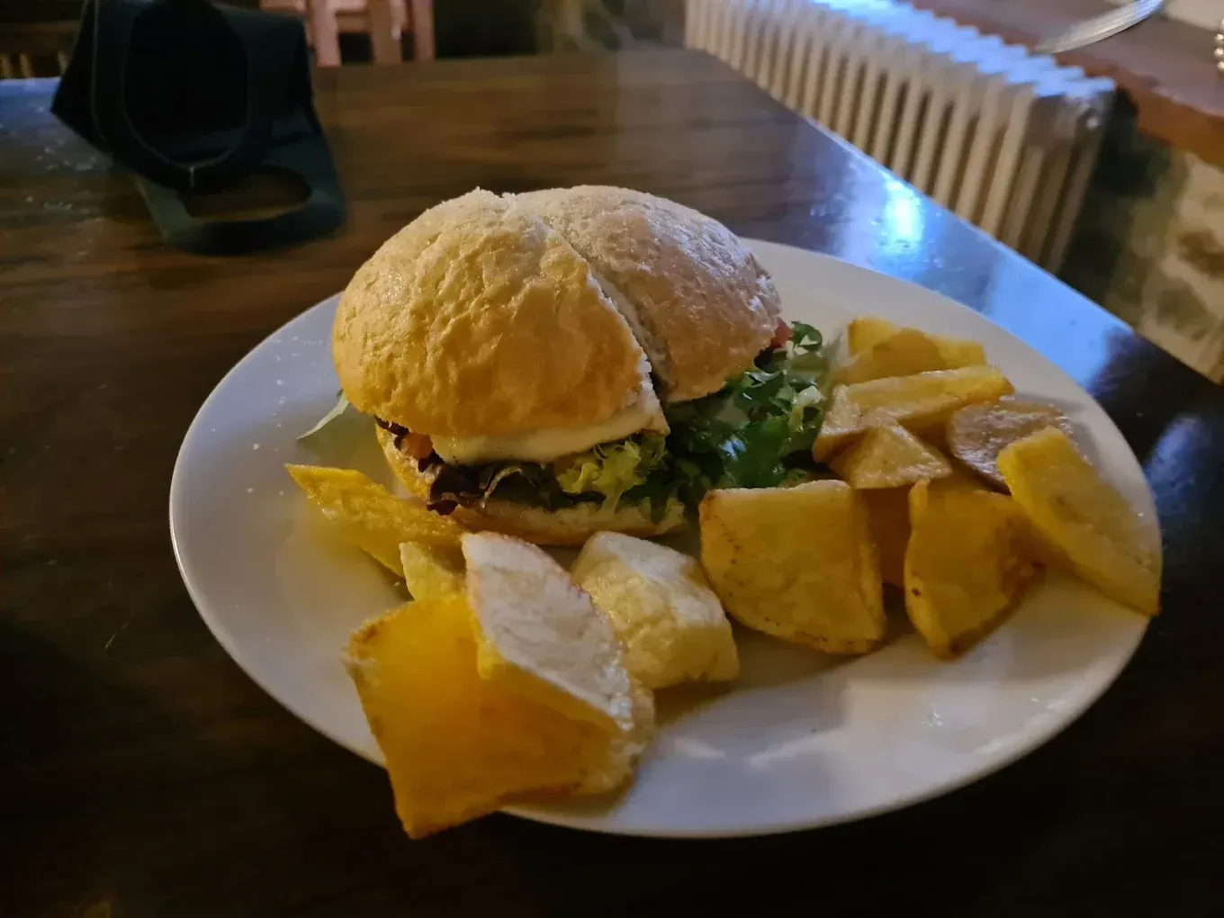 Hamburguesa y patatas fritas en un plato blanco sobre una mesa de madera, cerca de una cámara.