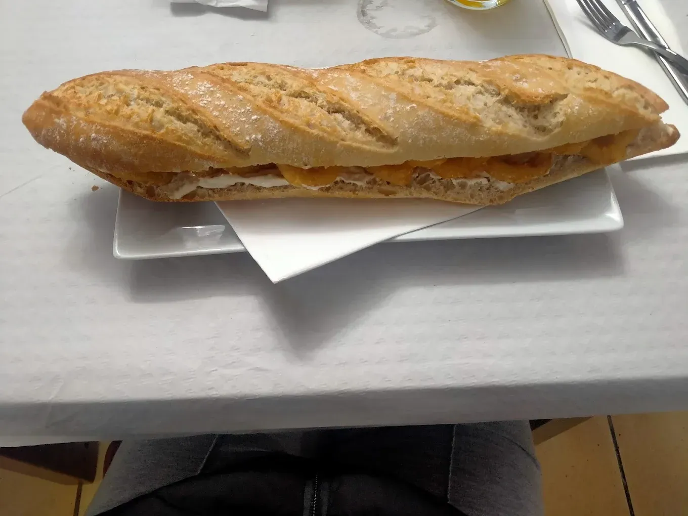 Sándwich en un plato blanco rectangular; pan baguette relleno. Servido sobre un mantel blanco.