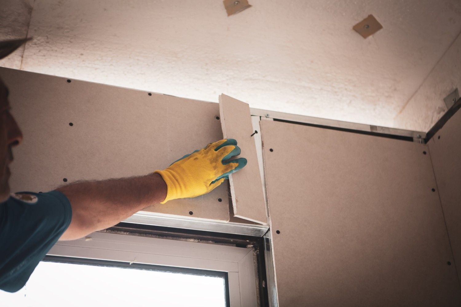 Persona con guantes amarillos instalando una esquinera de yeso cerca de una ventana. Paredes y techo beige.