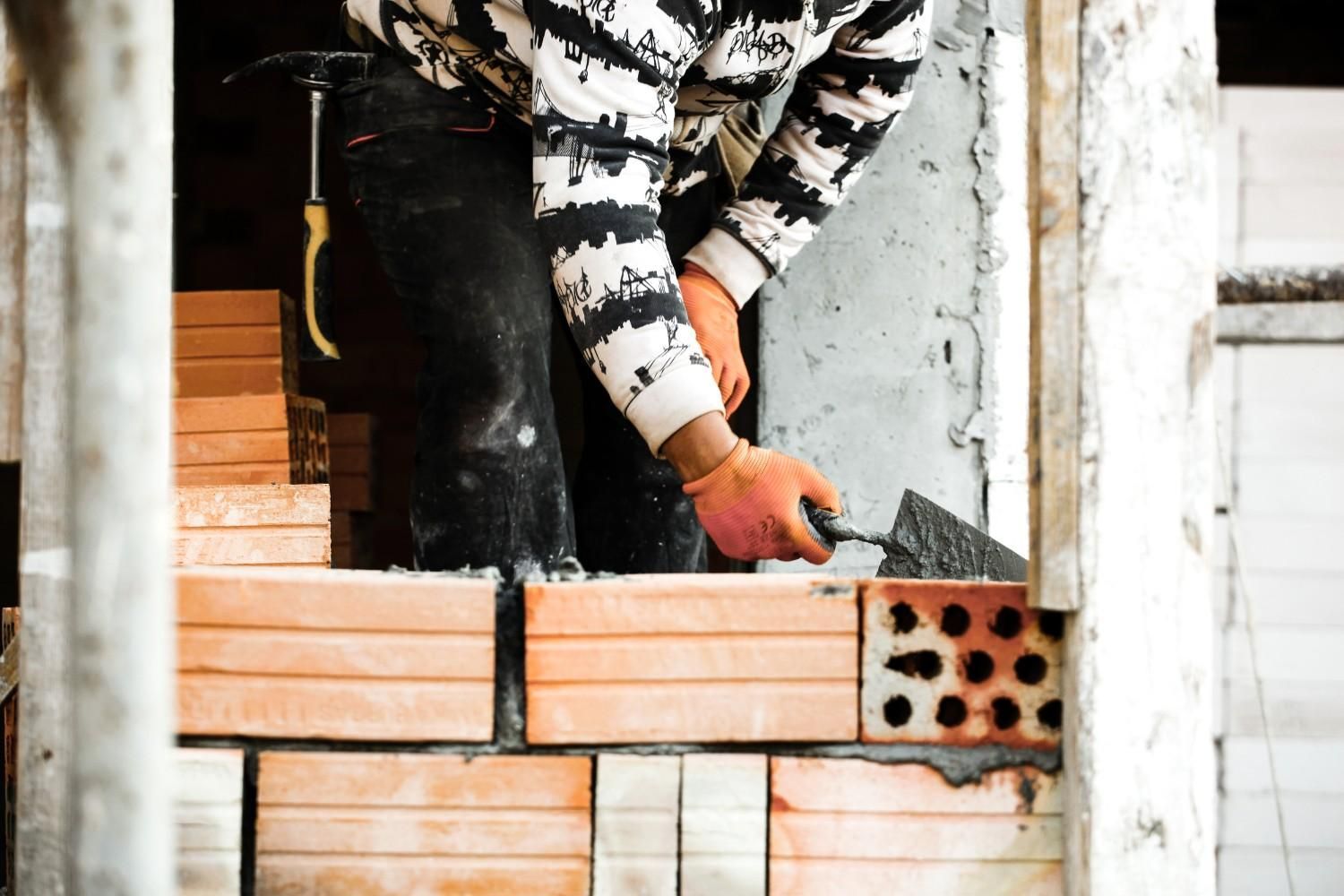 Albañil usando una paleta para colocar ladrillos en una pared, usando guantes de trabajo y un suéter blanco y negro.