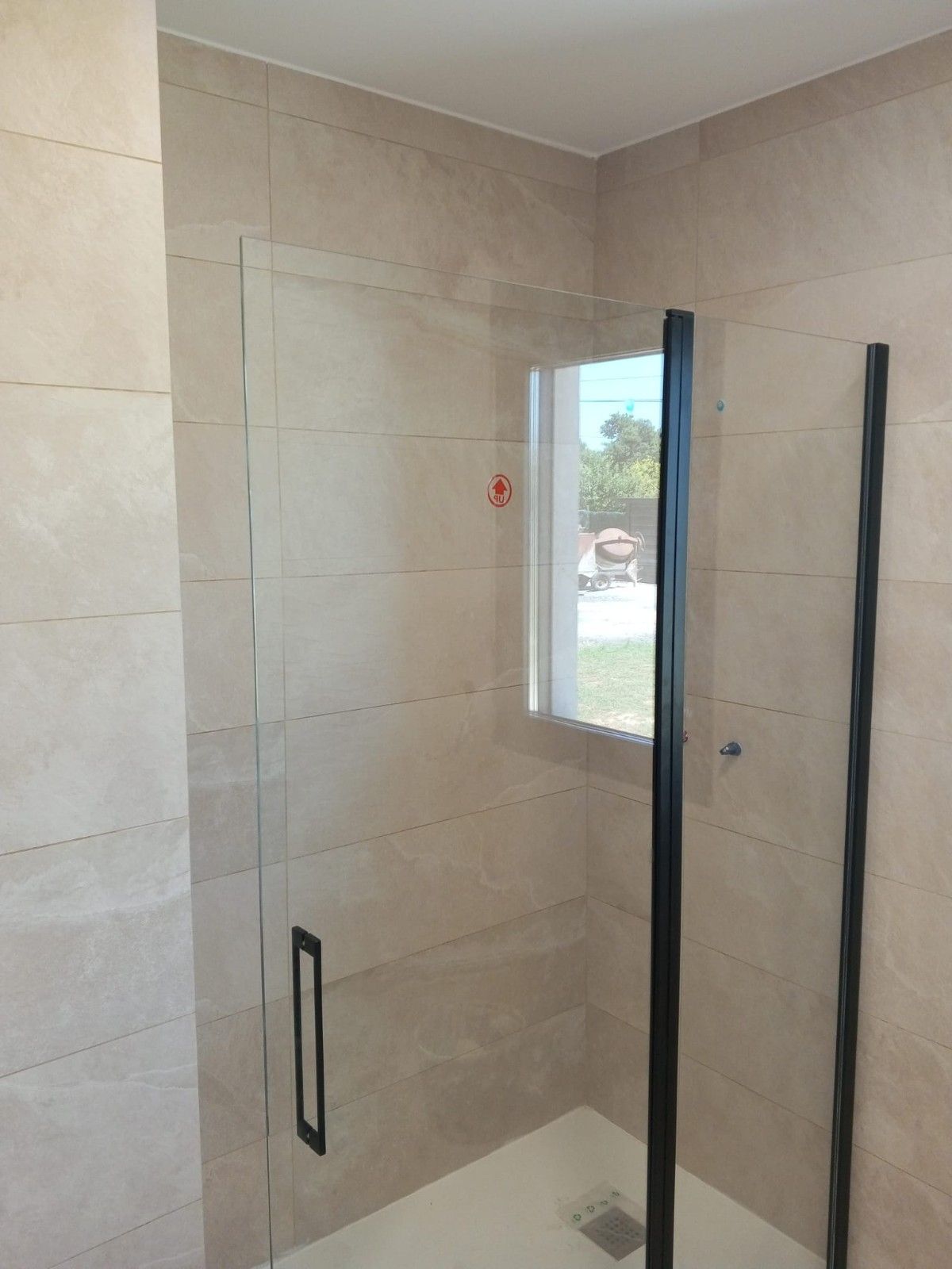 Cabina de ducha con puertas de cristal y marco de metal negro, paredes de azulejos de color tostado, ventana pequeña.