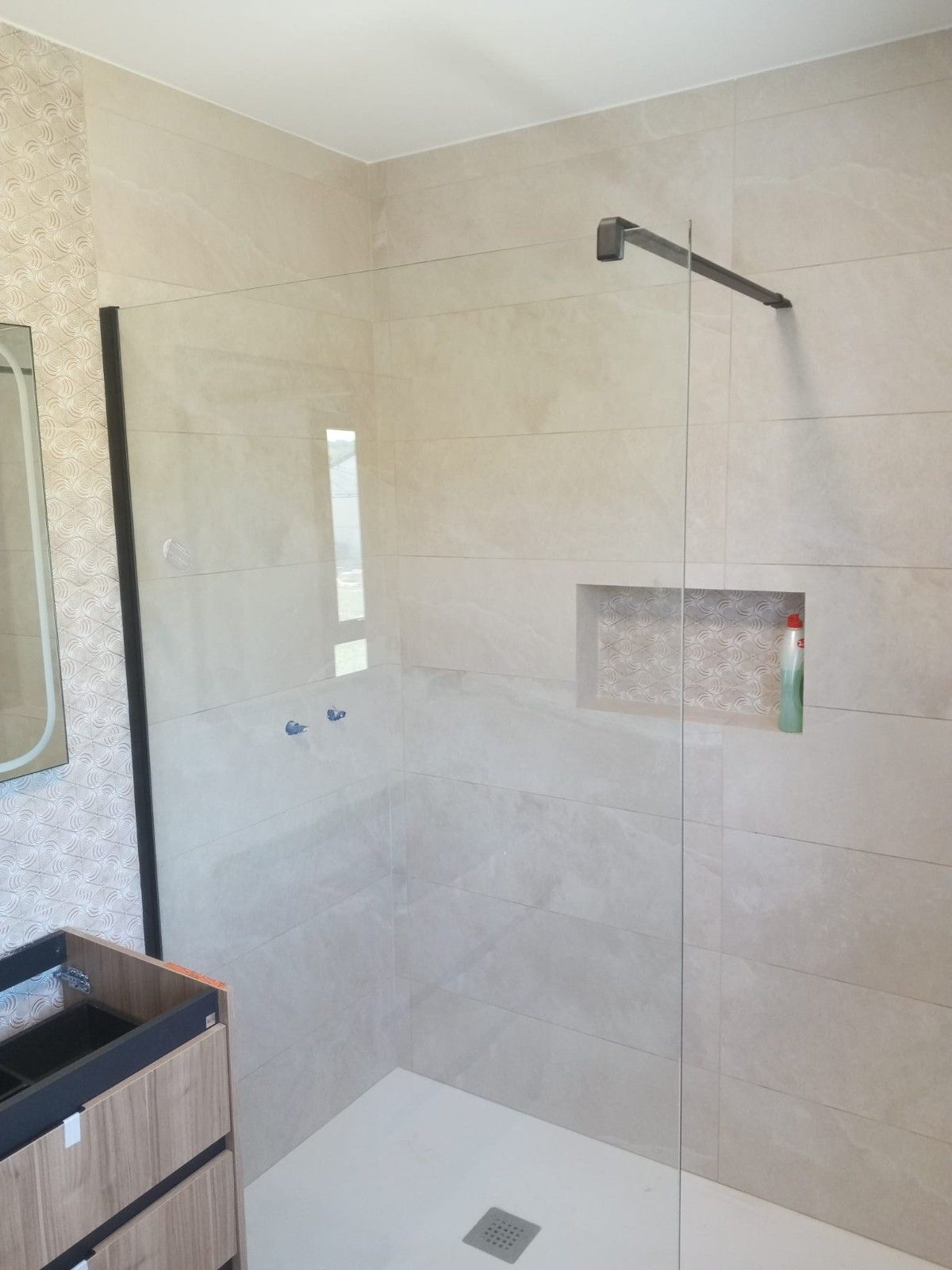 Ducha con mampara de cristal, paredes de azulejos claros y cabezal de ducha negro. El mueble se ve en la esquina inferior izquierda.