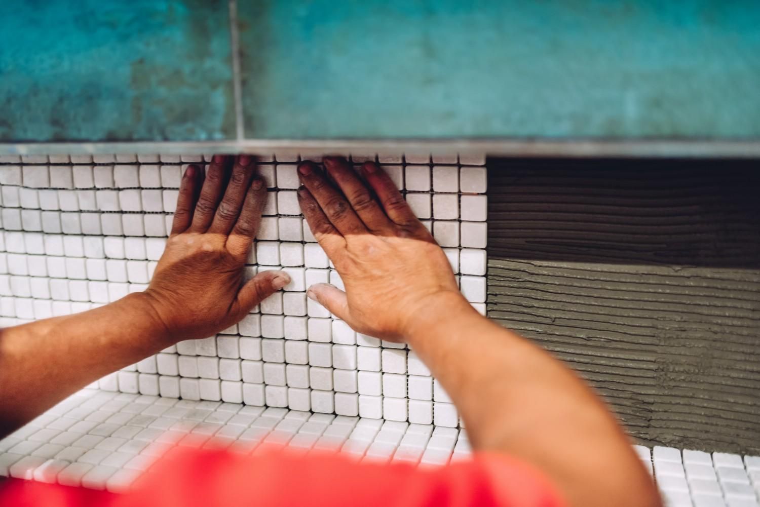 Manos de una persona instalando mosaicos en una pared. Adhesivo verde y pared azul visibles.